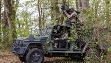 An ENOK AB light armored patrol vehicle is seen stationed in what appears to be a forest. The car is painted forest green, and two soldiers wearing green camouflage are seen crewing it. One is sitting in the driver's seat, while the other is on the back of the truck, aiming a high-millimeter rifle resting on his shoulder at something out of frame.